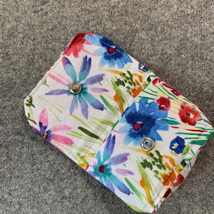 May include: A small white fabric pouch with a floral print in shades of blue, pink, yellow, and green. The pouch has a flap closure with a silver snap button.