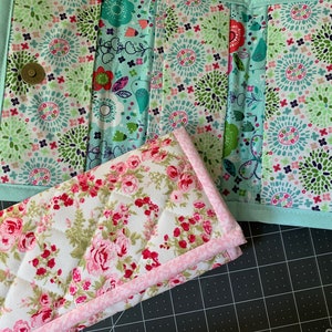 May include: Two quilted fabric wallets. One wallet features a floral pattern with pink roses on a white background, and the other has a floral design in shades of green and blue. Both have a light blue border.