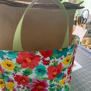 May include: A green canvas tote bag with floral print featuring red, pink, yellow, blue, and green flowers on a white background. The bag is filled with brown paper bags.