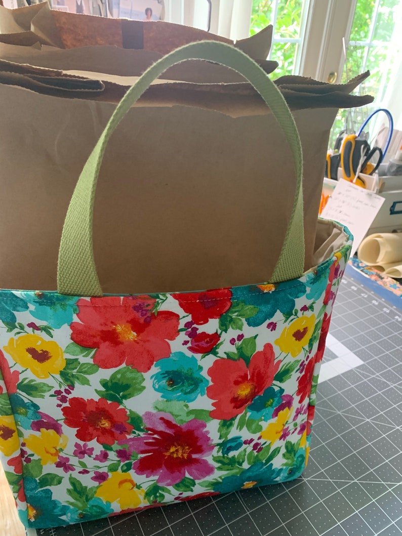 May include: A green canvas tote bag with floral print featuring red, pink, yellow, blue, and green flowers on a white background. The bag is filled with brown paper bags.