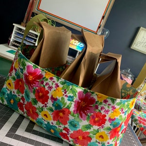 May include: A floral fabric storage bin with four brown paper bags inside. The bin is filled with bags and is sitting on a gray and black cutting mat.