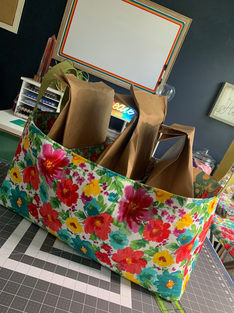 May include: A floral fabric storage bin with four brown paper bags inside. The bin is filled with bags and is sitting on a gray and black cutting mat.