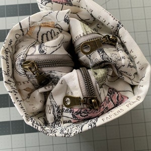May include: A white canvas zippered pouch with a floral and text print. The pouch has two zippers with brass pulls. The text on the pouch reads "PAPIERS PERKS".