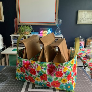 May include: A floral fabric storage bin with three brown paper bags inside. The bin has a green handle and is sitting on a black and white grid surface. The text "be brilliant." and "create" are visible in the background.