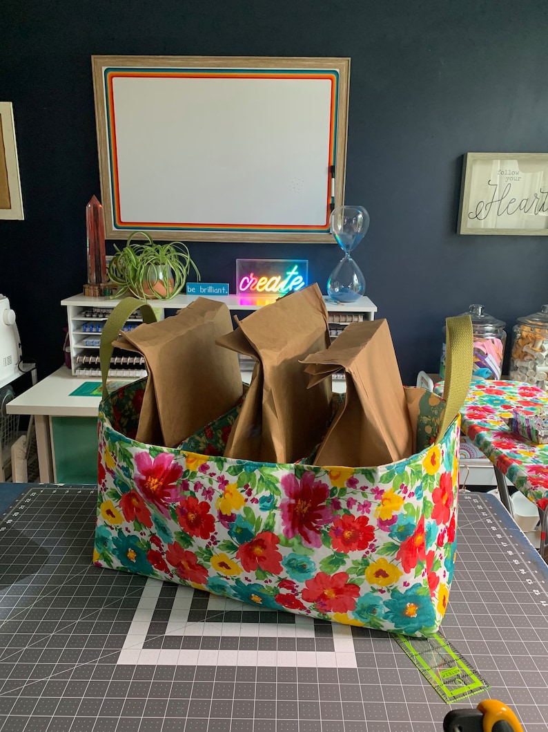 May include: A floral fabric storage bin with three brown paper bags inside. The bin has a green handle and is sitting on a black and white grid surface. The text "be brilliant." and "create" are visible in the background.