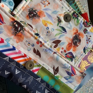 Puede incluir: Una bolsa de tela multicolor con un patrón floral y un cierre a presión. La bolsa está hecha de diferentes telas con patrones geométricos, incluyendo rayas, triángulos y lunares.
