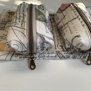 Peut inclure: Trois pochettes zippées avec un fond blanc et du texte noir et gris. Les pochettes ont un design vintage avec un motif floral et le texte "RISE", "TRAVAUX PUBLICS ET" et "25".