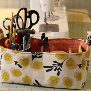 May include: A white fabric storage basket with yellow floral print and black accents. The basket has two compartments and two handles. It is filled with various craft supplies, including scissors, pens, and a hammer.