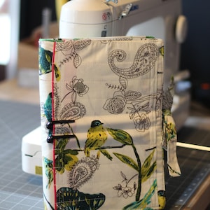 Peut inclure: Une pochette zippée en tissu blanc avec un motif floral et paisley. La pochette a une fermeture éclair noire et une bordure rose. La pochette est posée sur un tapis de coupe gris.