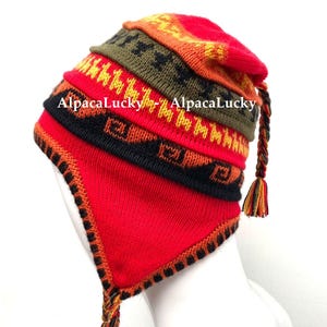 Può includere: Un cappello di maglia colorato con paraorecchie e nappa. Il cappello presenta strisce rosse, arancioni, gialle, nere e verdi con motivi geometrici. Il testo "AlpacaLucky" è stampato sul cappello.