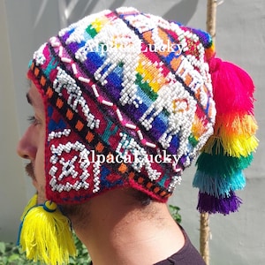Könnte beinhalten: Eine bunte Strickmütze mit weißen Perlen und Regenbogen-Quasten. Die Mütze hat ein geometrisches Muster und ist aus Alpakawolle gefertigt. Die Mütze wird von einer Person mit einem schwarzen Shirt getragen.