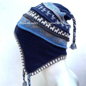 Könnte beinhalten: Eine blaue und graue Strickmütze mit einem langen geflochtenen Bommel. Die Mütze hat einen umklappbaren Ohrenschutz und ein Muster aus Sternen und Zickzacklinien. Der Text "AlpacaLucky" ist auf der Mütze sichtbar.