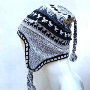 Könnte beinhalten: Eine grau, schwarz und weiß gestrickte Mütze mit Ohrenklappen und geflochtenen Quasten. Die Mütze hat geometrische Muster und den Text "AlpacaLucky". Die Mütze ist so konzipiert, dass sie Kopf und Ohren warm hält.