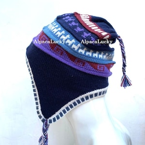 May include: A blue, purple, red and white knitted hat with a long braided tassel. The hat has a geometric pattern and the words "AlpacaLucky" are printed on the side.