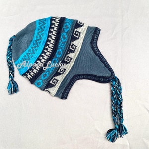 Può includere: Un cappello di lana blu e bianco con un motivo geometrico e due lunghi pon pon intrecciati. Il cappello ha una patta che copre le orecchie.