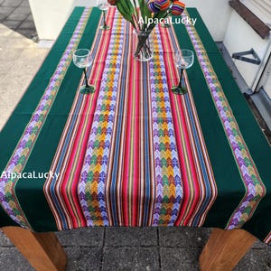 Op de afbeelding: Een donkergroen rechthoekig tafelkleed met een centrale loper met verticale strepen in rood, oranje, geel en blauw, geaccentueerd door ingewikkelde geometrische patronen. Een glazen vaas bevat stoffen tulpen in bijpassende kleuren en drie lege wijnglazen.