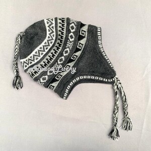 May include: Grey knitted hat with a white and black geometric pattern. The hat has two long, braided ties.