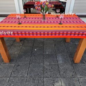 Op de afbeelding: Een rechthoekig tafelkleed met horizontale strepen in levendige tinten oranje, rood, roze en paars met geometrische patronen. De tekst AlpacaLucky is zichtbaar op de stof. Het kleed bedekt een houten tafel met vier doorzichtige glazen.