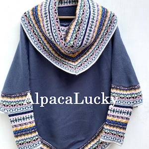 Op de afbeelding: Een blauwe alpaca wollen poncho met een wit, roze en geel geometrisch patroon rond de halslijn en mouwen. De poncho heeft een coltrui en hangt aan een houten hanger.