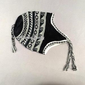 Könnte beinhalten: Eine schwarz-graue Strickmütze mit Ohrenklappen und geflochtenen Bändern. Die Mütze hat ein geometrisches Muster in Weiß und Grau. Der Text "Alpaca Lucky" ist auf der Mütze zu sehen. Die Mütze soll Kopf und Ohren warm halten.