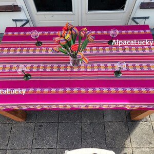 Peut inclure: Une nappe rectangulaire magenta vif avec des rayures horizontales jaunes, orange et violettes à motifs géométriques. Le texte AlpacaLucky apparaît deux fois sur le tissu. Un vase en verre transparent contient des tulipes en tissu colorées.