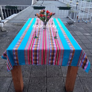 Op de afbeelding: Een rechthoekig tafelkleed in fel turquoise met een centraal verticaal paneel met veelkleurige strepen en geometrische patronen. De tekst AlpacaLucky is in het wit in het midden gedrukt. Een glazen vaas bevat verschillende kleurrijke stoffen tulpen.