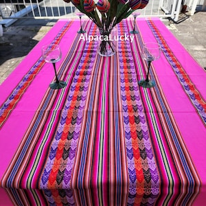 May include: A bright pink tablecloth featuring a central runner with vertical stripes in orange, purple, red, and black, decorated with geometric patterns. A glass vase holds fabric tulips in matching colours, and four wine glasses are placed on the table. The text AlpacaLucky is visible.
