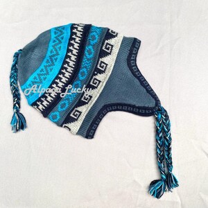 May include: A blue and white knitted hat with a geometric pattern and two long braided tassels. The hat has a flap that covers the ears.