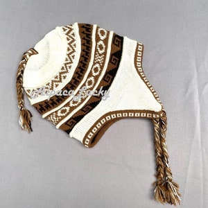 Könnte beinhalten: Eine weiße und braune Strickmütze mit geometrischem Muster und zwei langen geflochtenen Quasten.
