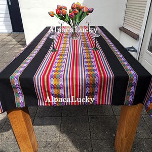 Op de afbeelding: Een rechthoekig tafelkleed met een effen zwarte rand en een centraal paneel met levendige, veelkleurige verticale strepen en geometrische patronen. De tekst AlpacaLucky staat twee keer op de stof gedrukt. Een glazen vaas met kleurrijke tulpen staat erop.