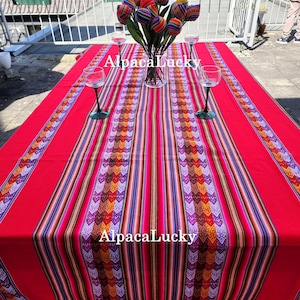 May include: A vibrant red rectangular tablecloth featuring a central panel of multicoloured striped and geometric patterns. The text AlpacaLucky is printed twice on the fabric. A clear glass vase holds several colourful fabric tulips.