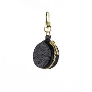 May include: A black, round, leather keychain pouch with a gold-coloured zip and clasp. The pouch has a textured surface and is designed to hold small items. The keychain is suspended against a white background.