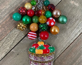 May include: A red, green, and gold beaded keychain with a charm of a basket filled with red, green, and orange apples. The charm is covered in rhinestones. The keychain is labeled "Tushka Designs".