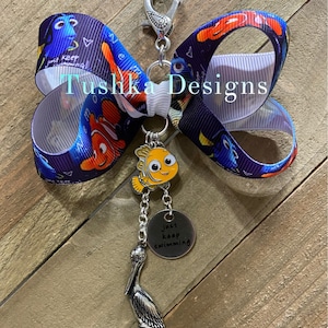 Op de afbeelding: Een blauwe en witte strik met een Finding Nemo-thema. De strik heeft een zilveren bedel met een pelikaan en een bedel met de tekst "Just Keep Swimming".