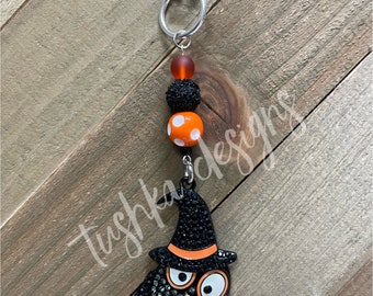 Op de afbeelding: Een zwarte uilbedel met oranje en witte stippen en een zwarte hoed. De bedel is bevestigd aan een zilveren ketting met een karabijnhaak.