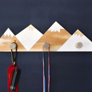 Puede incluir: Un gancho de pared de madera con forma de montaña con tres ganchos, pintado de blanco y marrón con un diseño de cordillera. Los ganchos son de madera y tienen un acabado natural. Hay tres artículos colgados de los ganchos, incluyendo una cinta roja y dos cintas azul y blanca.