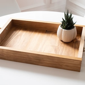 Puede incluir: Una bandeja de madera con asas doradas y una pequeña planta suculenta en maceta. La bandeja está hecha de madera natural y tiene un aspecto rústico.
