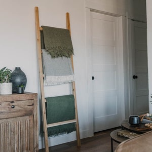 Puede incluir: Una escalera de madera apoyada en una pared blanca con tres mantas verdes, grises y blancas dobladas en los peldaños.