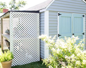 May include: A gray shed with white trim and light blue double doors. The shed has a white lattice fence attached to the side. A green bush with white flowers is in front of the shed.