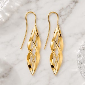 Könnte beinhalten: Ein Paar goldfarbene Ohrhänger mit einem länglichen, durchbrochenen Design aus mehreren ineinander verschlungenen, gedrehten Metallbändern, die eine käfigartige Struktur bilden. Die Ohrringe hängen an einfachen, geschwungenen Ohrhaken.