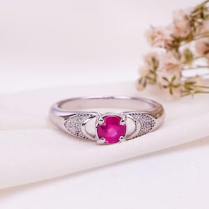 Könnte beinhalten: Ein silberner Ring mit einem roten Rubin-Edelstein und kleinen klaren Edelsteinen auf der Ringschiene.