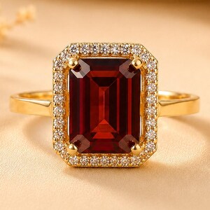 Könnte beinhalten: Ein Goldring mit einem großen, rechteckigen, tiefroten Edelstein im Smaragdschliff in der Mitte. Der zentrale Stein ist von einem Kranz aus kleinen, runden, klaren weißen Steinen in einer Goldfassung umgeben, die auf einer schlichten Goldschiene sitzt.