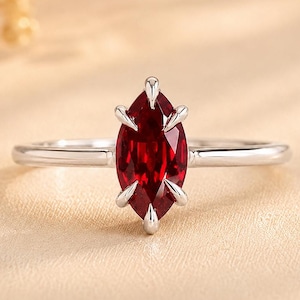 Könnte beinhalten: Ein silberfarbener Ring mit einem einzelnen, tiefroten Edelstein im Marquise-Schliff, der in einer Fassung mit sechs Krappen sitzt. Die Ringschiene ist schmal und poliert und bietet ein klares Metallfinish, das den leuchtenden Stein betont.
