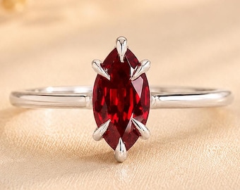 Op de afbeelding: Een zilverkleurige ring met een enkele dieprode edelsteen in marquise-slijpvorm, gezet in een zetting met zes pootjes. De ringband is dun en gepolijst, met een strakke metalen afwerking die de levendige, gefacetteerde steen in het midden accentueert.