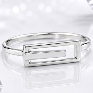 Könnte beinhalten: Ein polierter silberfarbener Ring mit einem rechteckigen, offenen Rahmendesign und einem horizontalen Balken in der Mitte. Die Ringschiene ist dünn und glatt und reflektiert das Licht auf einer hellen Oberfläche mit verstreuten Kristallakzenten.