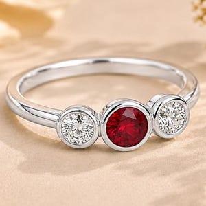 Könnte beinhalten: Ein silberfarbener Ring mit einem zentralen, rund geschliffenen roten Edelstein, der von zwei kleineren, rund geschliffenen klaren weißen Edelsteinen flankiert wird. Jeder Stein ist in einer glatten Zargenfassung auf der polierten Ringschiene gefasst.