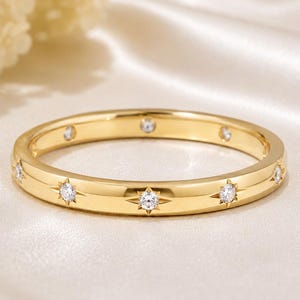Könnte beinhalten: Ein goldener Ring mit einer Reihe von gleichmäßig verteilten, klaren, rund geschliffenen Steinen. Jeder Stein ist in eine sternförmige Gravur auf der polierten Goldoberfläche eingefasst, was ein sich wiederholendes Muster ergibt.
