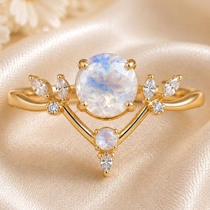 Könnte beinhalten: Ein goldener Ring mit einem großen, runden, schillernden Mondstein in der Mitte, akzentuiert durch kleinere runde Mondsteine und klare Steine im Marquise-Schliff, die in einem V-förmigen, blattartigen Muster entlang der Ringschiene angeordnet sind.