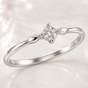 Könnte beinhalten: Ein silberfarbener Ring mit einem einzelnen, rund geschliffenen, klaren Stein in einer Vier-Krappen-Fassung. Die Ringschiene ist auf beiden Seiten des zentralen Steins mit zwei kleinen, glatten, blattförmigen Metallelementen verziert.
