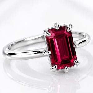 Könnte beinhalten: Ein silberfarbener Ring mit einem rechteckigen, im Smaragdschliff geschliffenen, tiefroten Edelstein in einer Krappenfassung. Die Ringschiene ist glatt und poliert und zeigt ein klares Metallfinish, das die lebendige, satte Farbe des Steins ergänzt.
